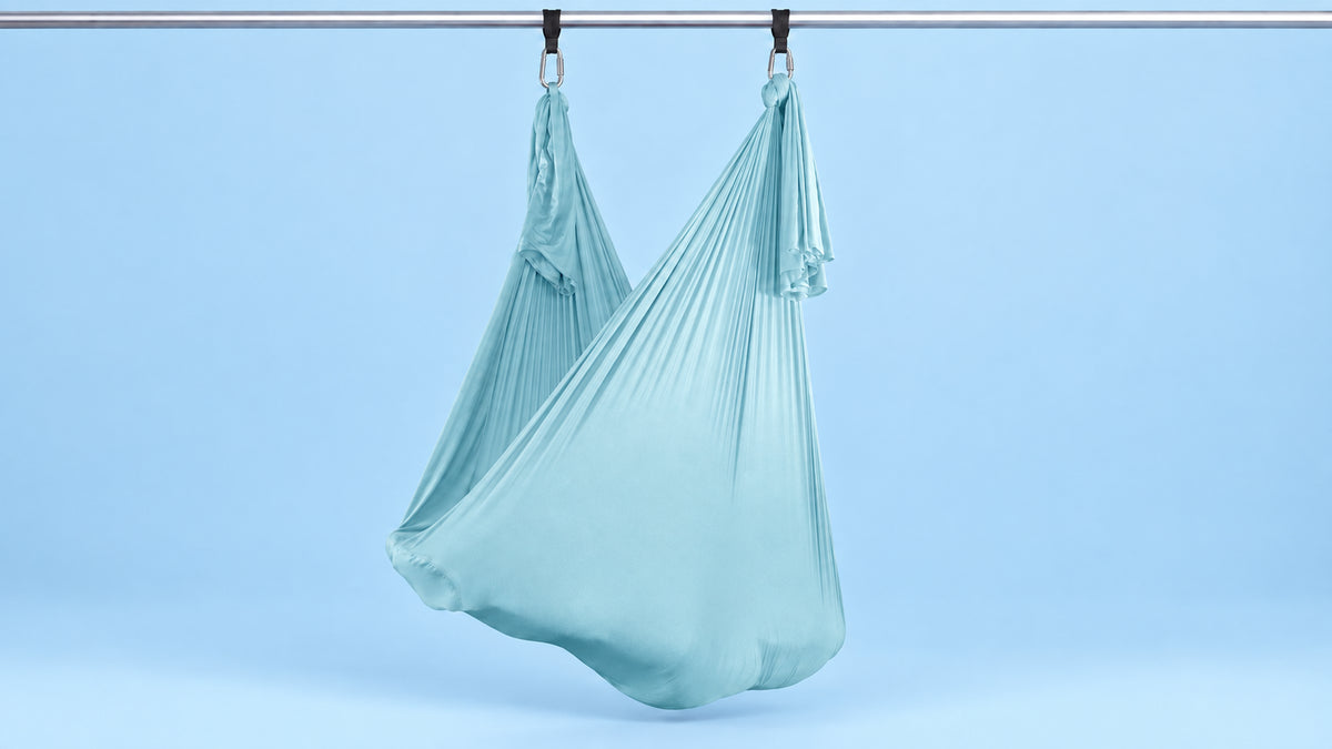 Hellblaues Aerial Yoga Tuch an zwei Punkten als Hängematte aufgehängt vor blauem Hintergrund